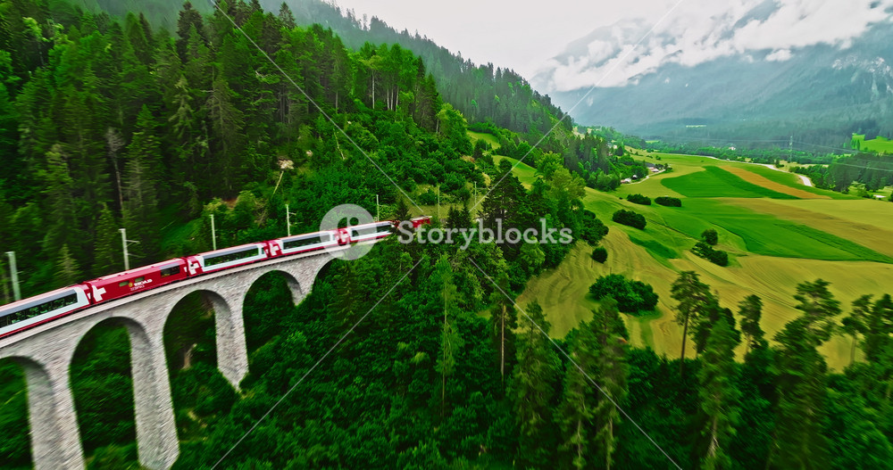 Aerial video of the red train traversing the Landwasser viaduct. The Swiss Express travels through the green meadows and mountains of