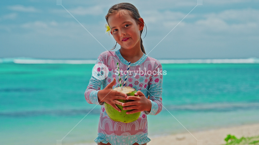 A close-up portrait of little girl drinking a fresh coconut cocktail with turquoise water on the shore of the Indian Ocean. A smile on the