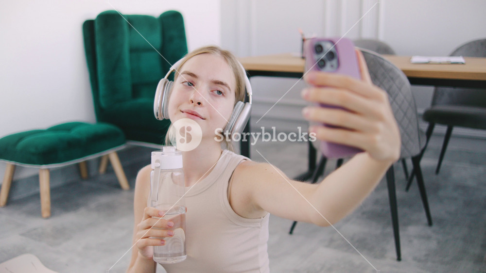 Young yoga sport fitness woman takes a selfie while sitting on the floor of a stylish room with headphones and a water bottle