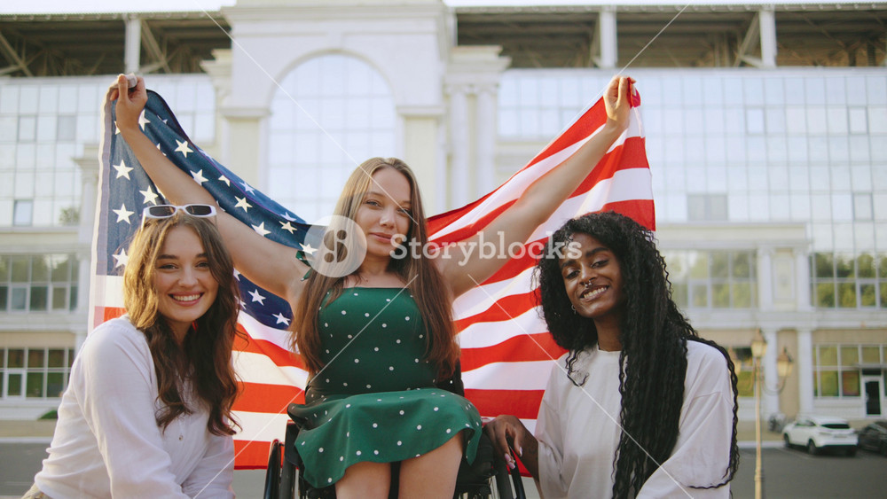 three female multiracial friends one inclusive woman in a wheelchair celebrate together while holding an American flag on a sunny day at a