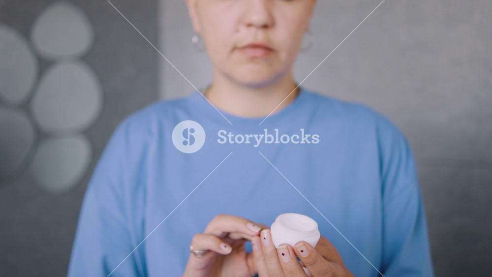 A bald woman, a cancer survivor, opens a white jar of facial cream in the bathroom, applies it to her face while looking at the camera as if was the mirror.