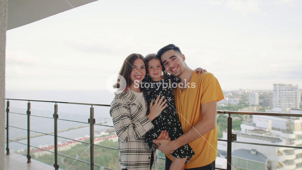 Cheerful family with little daughter enjoys a sunny day on a balcony overlooking the city and ocean with smiles and playful expressions