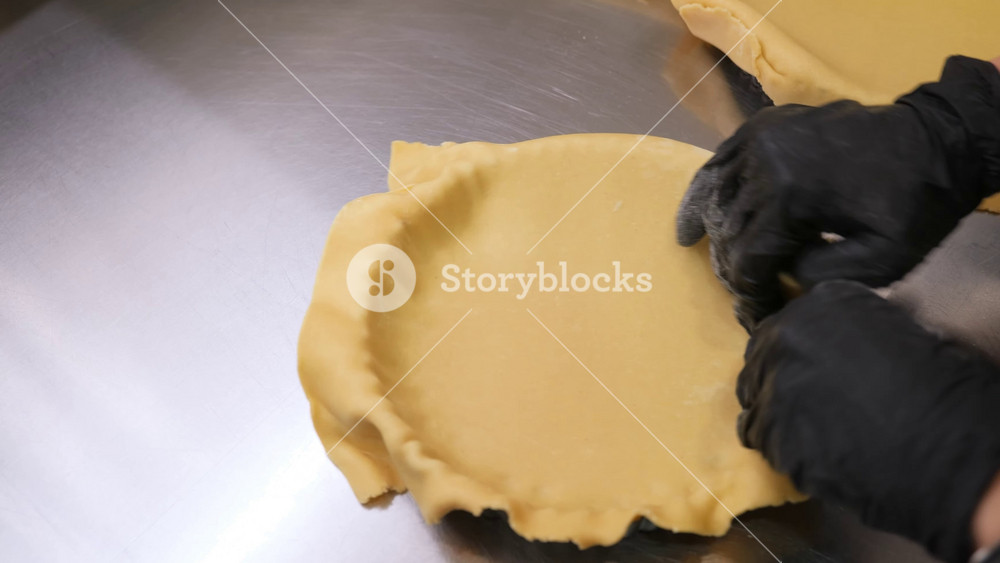Hands in black gloves carefully placing and smoothing a sheet of yellow sweet pastry dough into a pie tin on a professional stainless steel kitchen counter.