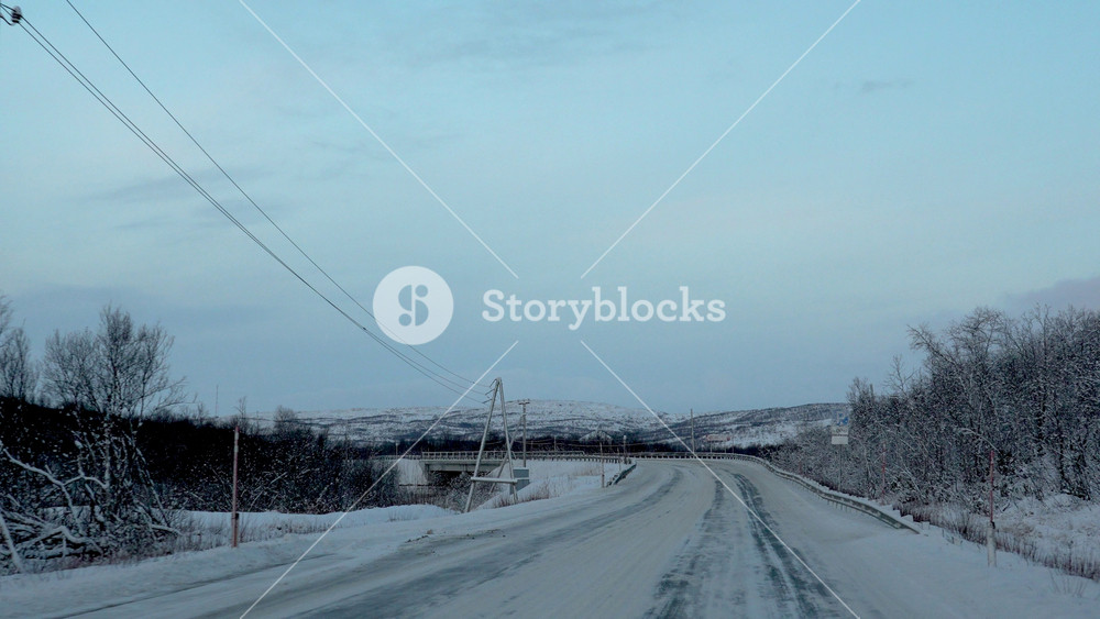 A perspective shot of an icy and snow-covered road passing through a landscape of snowy trees and scrub, with power lines and a small bridge in the distance