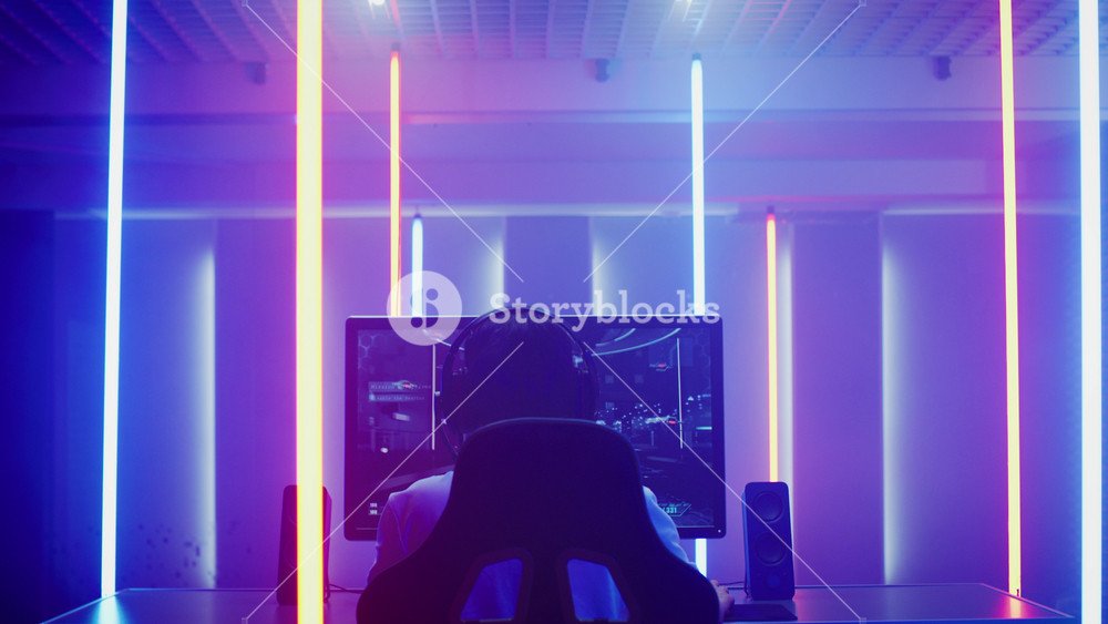 Back View Shot of the Professional Gamer Playing in First-Person Shooter Online Video Game on His Personal Computer. Room Lit by Neon Lights in Retro Arcade Style. Online Cyber e-Sport Internet Championship.