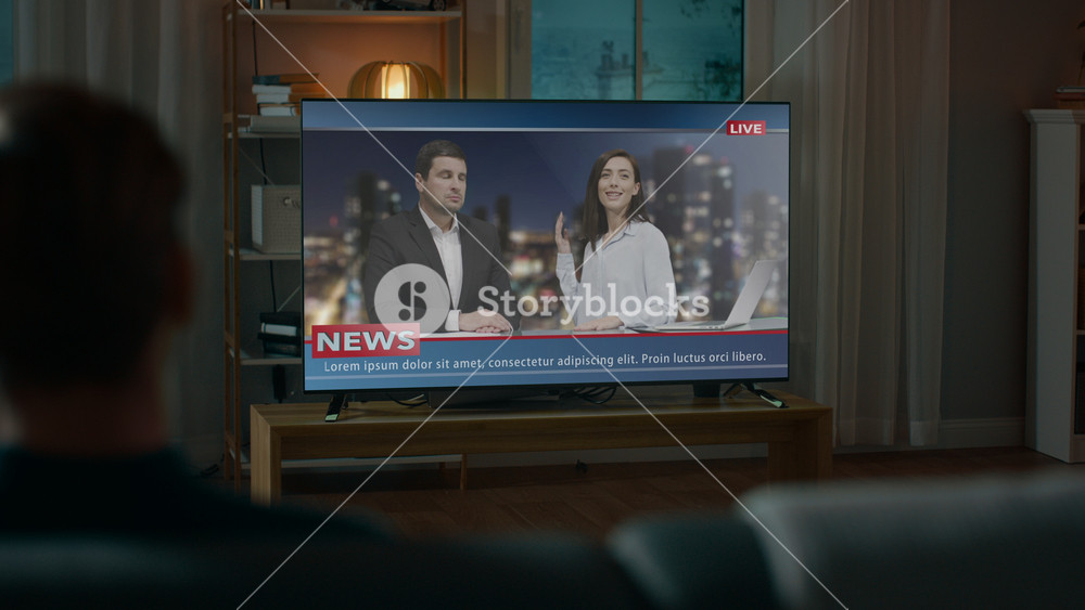 Man Watches Late Night News Show on TV while Sitting on a Couch at Home in the Evening. Two Presenters Talk and Joke on TV. Cozy Living Room with Warm Lights. Over the Shoulder Shot.