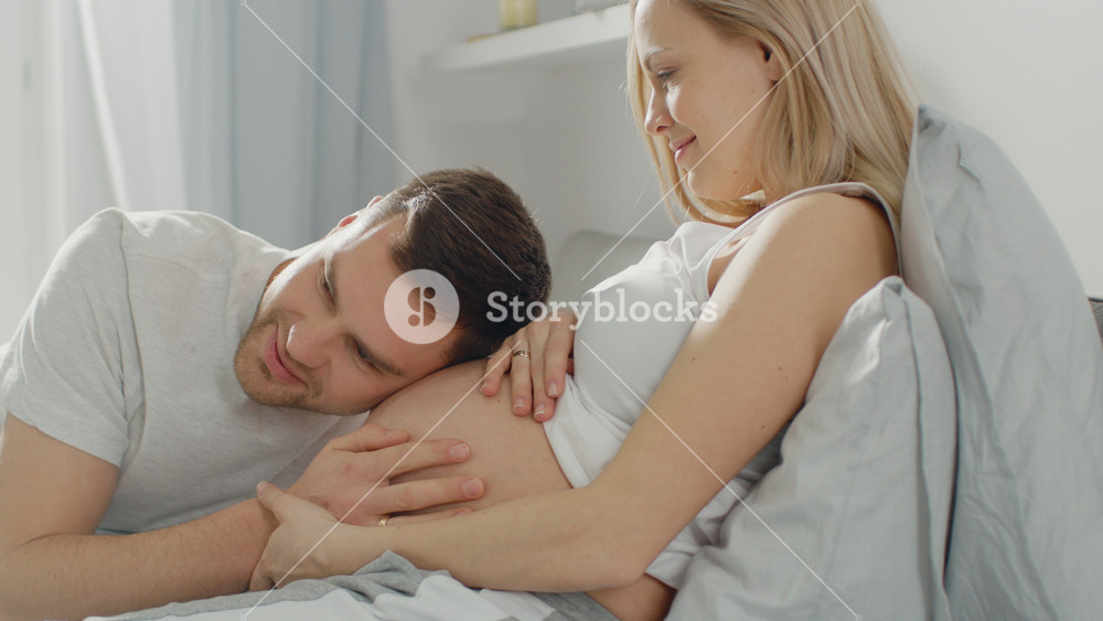 Happy Young Couple Cuddling Together in the Bed, Young Woman is Pregnant, Loving Partner Leans and Listens to a Baby Moving and it's Heartbeat in the Belly. Tender and Loving Moments of Family Life.