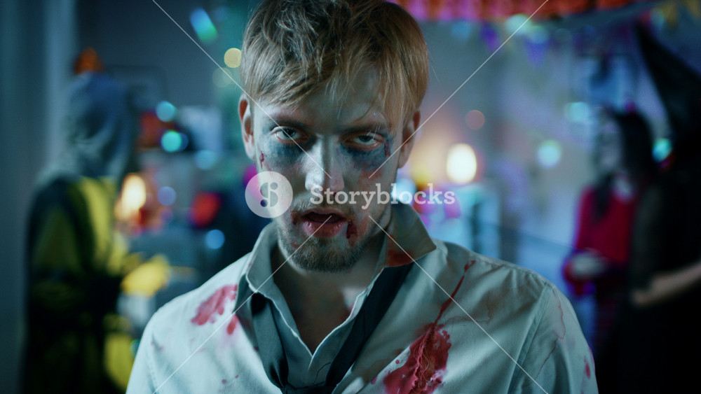 Halloween Costume Party: Portrait of Brain Dead Zombie Wearing Bloody Suit Smiles Creepily. In the Background Monsters Having Fun and Dancing in the Decorated Room