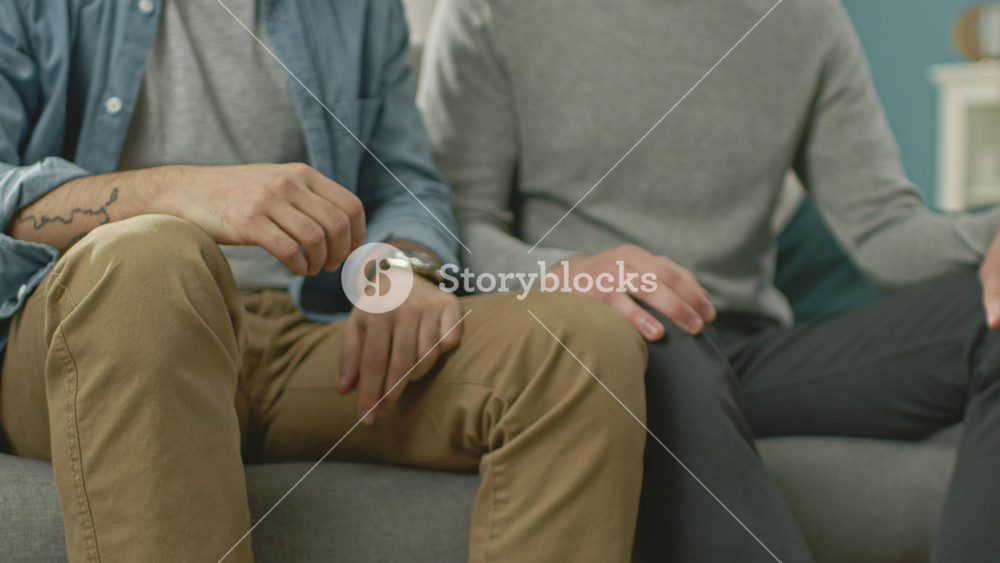 Close Up of a Gentle Male Gay Couple Sitting Together on a Sofa at Home. Boyfriend Puts His Hand on Partner's. They are Casually Dressed and Their Room Has Modern Interior.