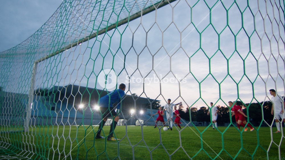 On Soccer Championship Player Kicks the Ball and Goalkeeper Tries to Defend Goals but Jumps and Fails to Catch the Ball. Camera Shot from Behind the Net with whole Stadium Visible.