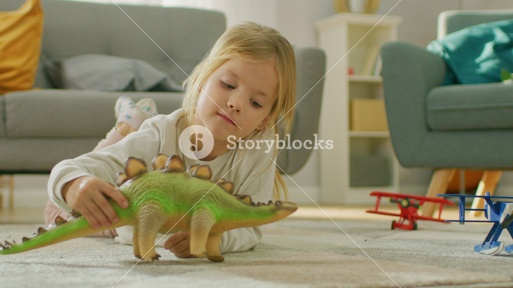 Cute Little Girl Blond Laying on a Carpet at Home, Plays with Toy Dinosaurs and Airplanes. Happy Child Playing with Toys in Sunny Living Room.