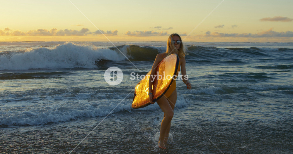Beautiful Young Woman in the Swimsuit Walks out of the Sea while Carrying Surfboard. She is Looks Gorgeous and Slim, Has Blonde Hair. Big Oceanic Waves and Sunset in the Background.
