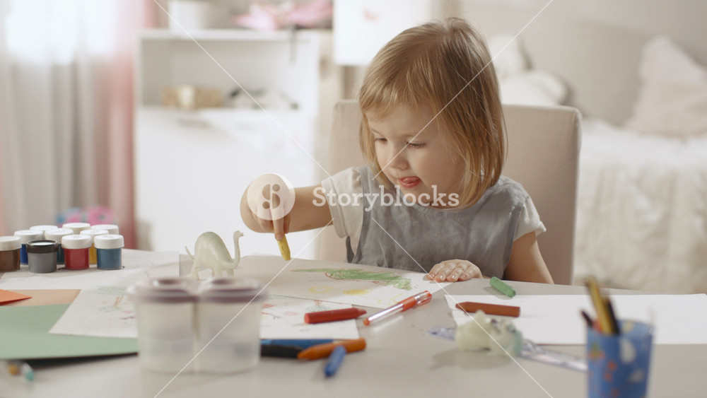 Cute Little Girl Sits at Her Table and Draws with Crayons. Her Room Is Pink, Pretty Drawings Hanging on the Walls, Many Toys Lying Around.