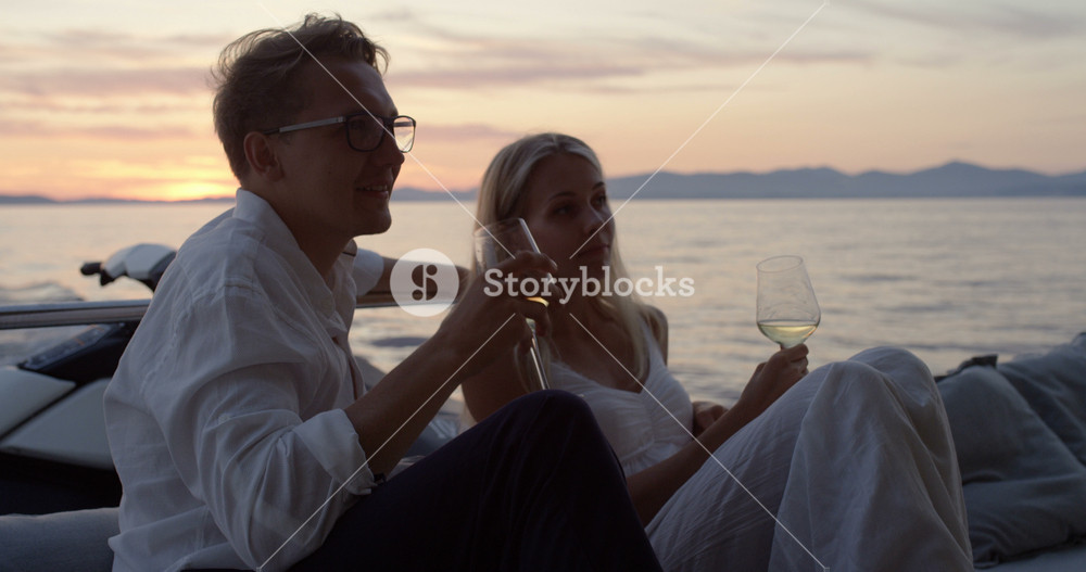 Young Couple Talk, Drink Champagne in the Stern of the Moving Yacht. They Have Great Romantic Evening. In the Background Island with Small Village.
