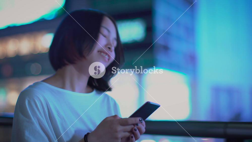Portrait of the Attractive Alternative Japanese Girl Wearing Casual Clothes Uses Smartphone, Walks Through the City. In the Background Big City Advertising Billboards Lights Glow in the Night.