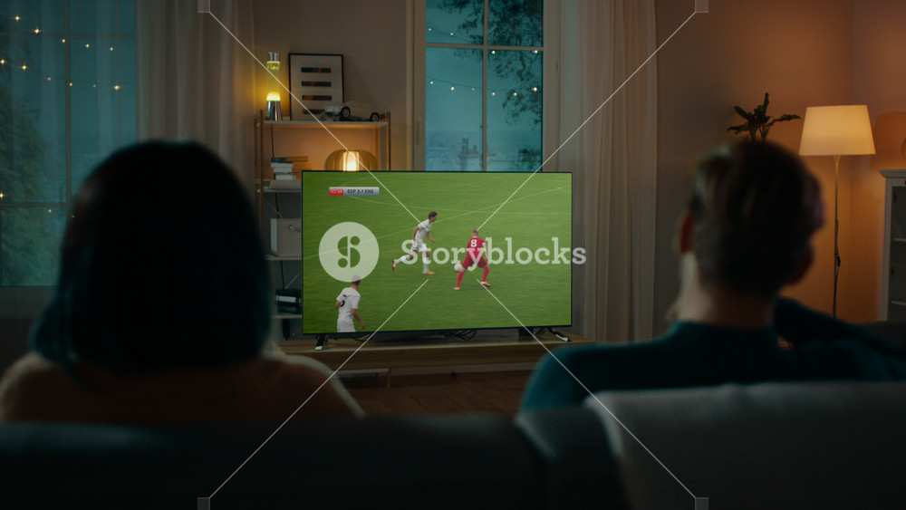 Couple Watches Professional Soccer Match on TV while Sitting on a Couch at Home in the Evening. Boyfriend and Girlfriend Football Fans Watch Sports Even. Back View Shot.