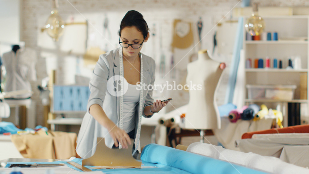 Beautiful Brunette fashion, designer, Lays Out Templates on the fabrics and Consults Her Smartphone. She Works in a Light Colorful Studio Full of Various Clothes, Fabrics and Sketches on the Wall.