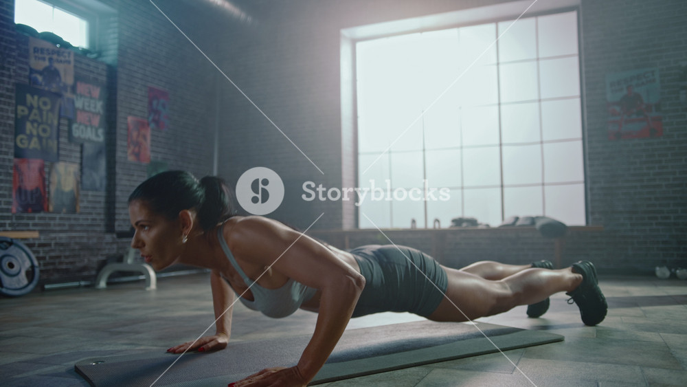 Strong and Fit Athletic Woman in Sport Top and Shorts is Doing Push Up Exercises in a Loft Style Industrial Gym with Motivational Posters. It's Part of Her Cross Fitness Training Workout.