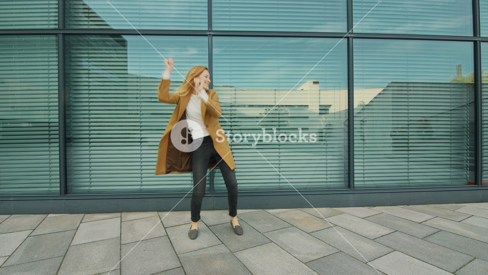 Cheerful and Happy Young Woman Actively Dancing in Front of a Glass Building. She's Wearing a Long Brown Coat. Scene Shot Next to Business Center. Day is Bright.