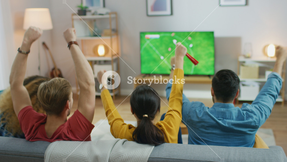At Home Diverse Group of Sports Fans Sitting on a Couch Watching Important Soccer Match on TV, They Cheer for the Team, Celebrate Victory after Team Scoring Winning Goal. Cozy Room with Snacks and Drinks.