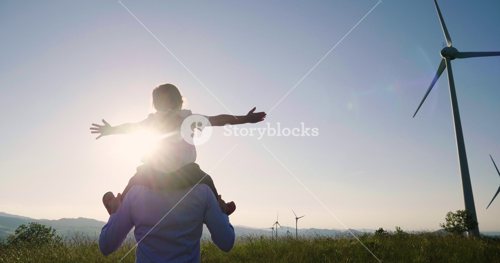 A Caucasian Man Carries a Joyful Child on His Shoulders, Arms Outstretched, Amidst a Serene Wind Farm Landscape, Symbolizing Hope and the Promise of Sustainable Energy for Future Generations.