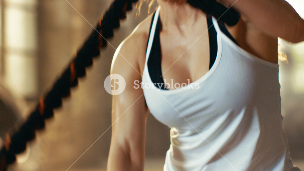 Close-up of a Athletic Female in a Gym Exercising with Battle Ropes During Her Cross Fitness Workout/ High-Intensity Interval Training. She's Muscular and Sweaty, Gym is in Industrial Building.
