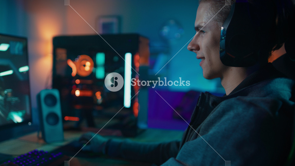 Happy Gamer in Headset with a Mic Playing Shooter Online Video Game on His Personal Computer. He Gives Commands to Other Players. Room and PC have Colorful Neon Led Lights. Cozy Evening at Home.