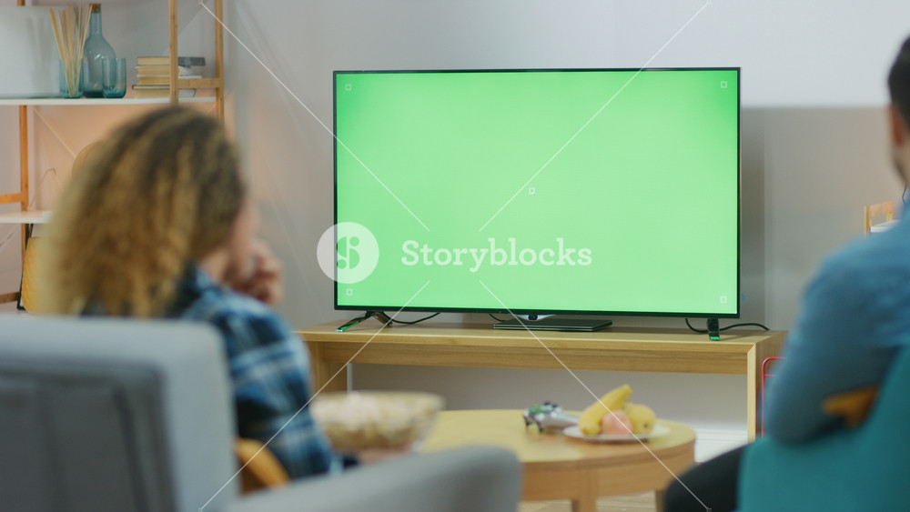 Happy Couple Sitting At Home in the Living Room Watching Green Chroma Key Screen TV, Relaxing on a Couch. Couple Room Watching Sports Match, News, Show or a Movie.