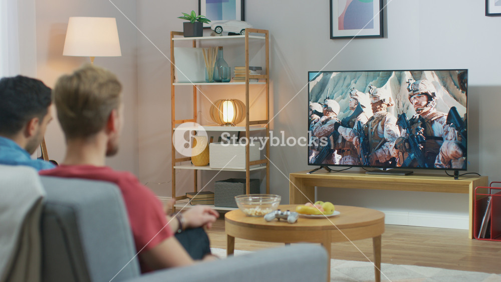 Over the shoulder view of Two Friends Relaxing on a Couch Watching War Movie on a TV In Living Room. Modern Military Warfare Action with Shooting and Explosions Shown on a Television.