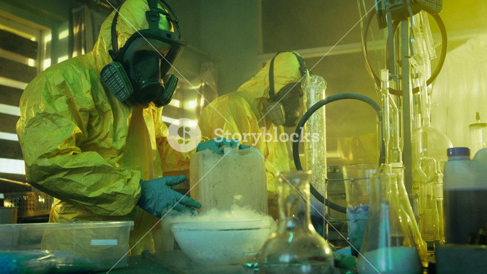 In the Underground Drug Laboratory Two Clandestine Chemists Covered in Protective Coveralls and Gas Masks Mix Chemicals to Synthesise Drugs. They Work in the Abandoned Building.