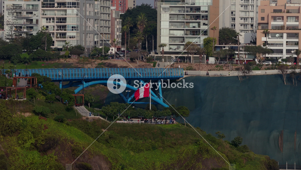 Aerial view of the Puente de la Paz pedestrian bridge with a waving Peruvian flag, surrounded by the green cliffs of the Costa Verde