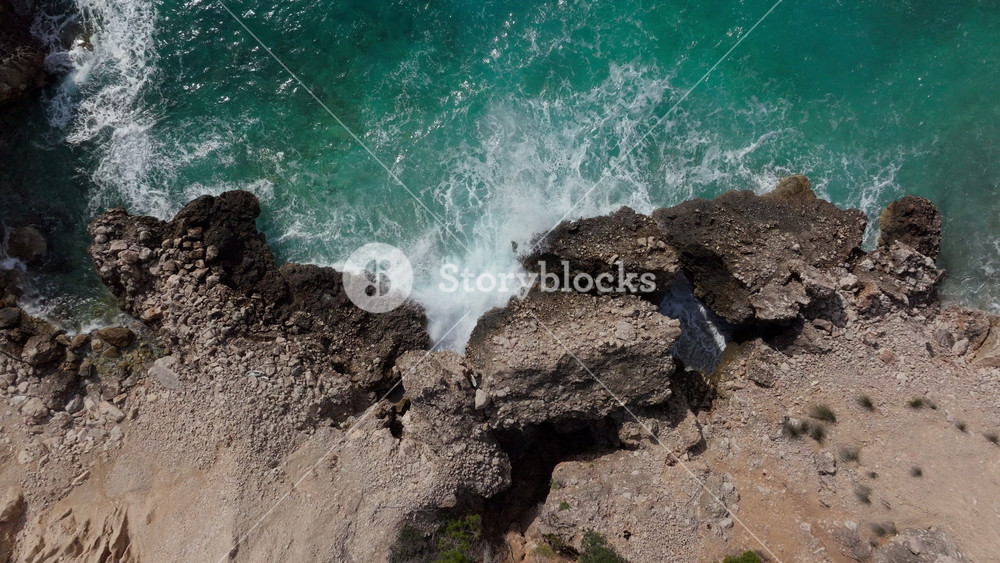 Scenic aerial top down view of beautiful turquoise sea waves splashing against a rugged, rocky coastline on the island of Mallorca, Spain