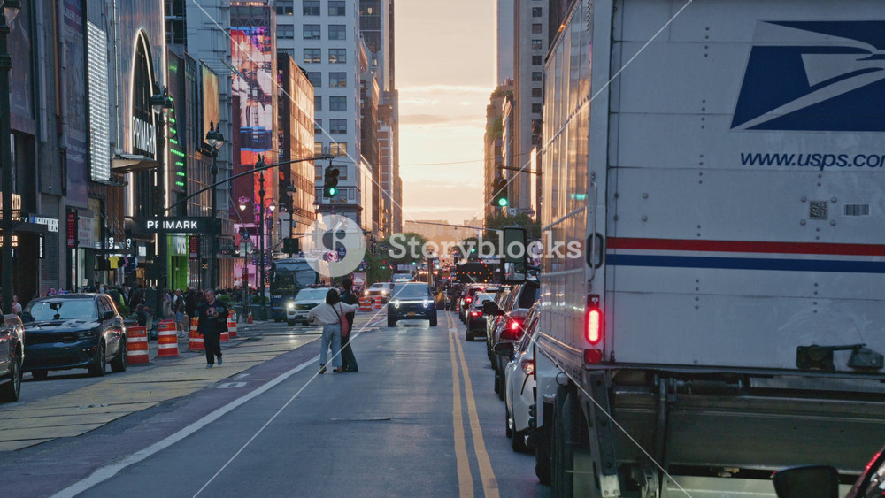 New York City street with pedestrians crossing at sunset and heavy traffic including USPS truck, cars driving and stopping at traffic light