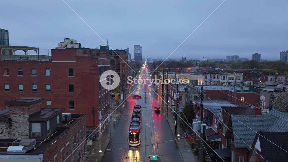 Streetcar traveling on wet city street during rainy twilight in downtown area with traffic and building lights creating reflections on