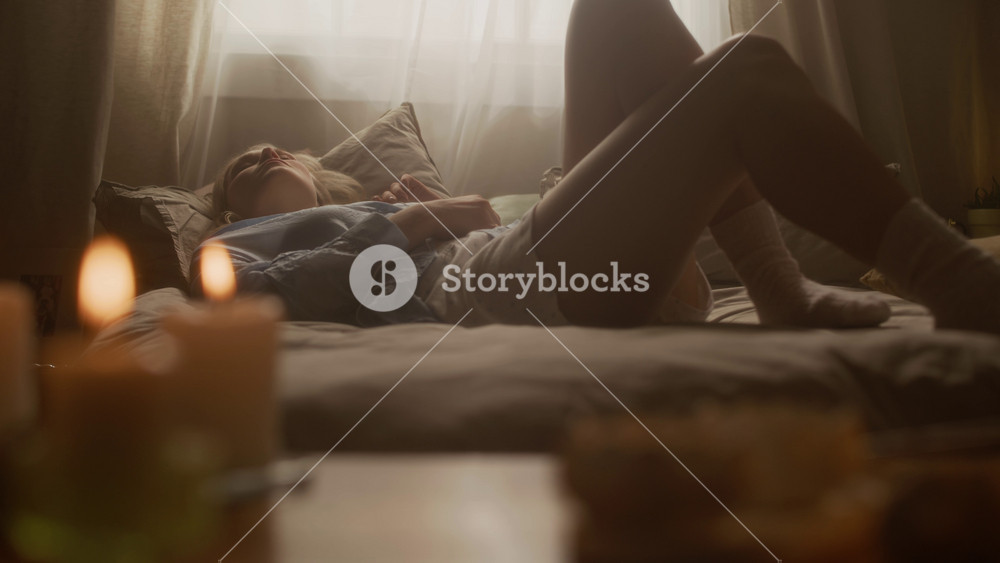 Handheld slow motion of relaxed Caucasian woman lying in bed, listening to music and moving foot to beat in room with warm lighting and lit candles