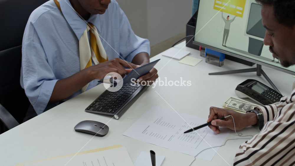 Handheld shot of young African American bank clerk in formalwear examining passport of male client reading document at desk with paper banknotes and terminal for contactless payment