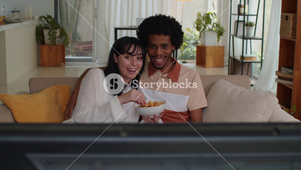 Medium long shot of young emotional biracial couple watching movie on TV and eating popcorn when sitting together on couch at Valentines Day at home