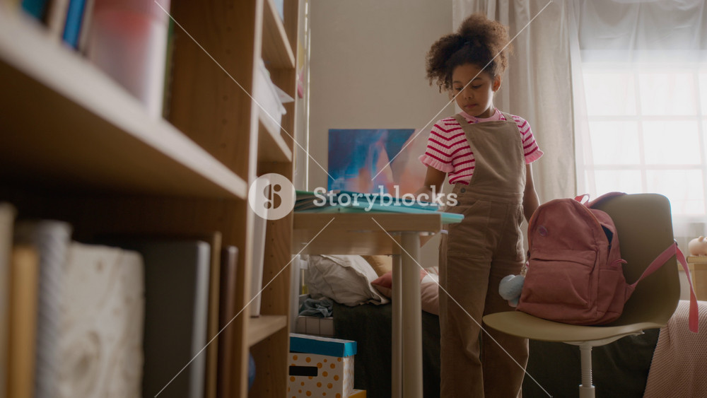 African American child packing school supplies into pink backpack in bedroom, preparing for school day. Ideal for back-to-school marketing campaigns, parenting promos, and educational content