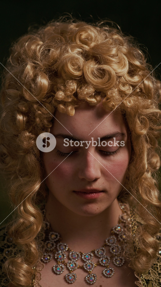 Vertical close up portrait of serious young woman wearing baroque curly blonde wig and necklace looking at camera indoors
