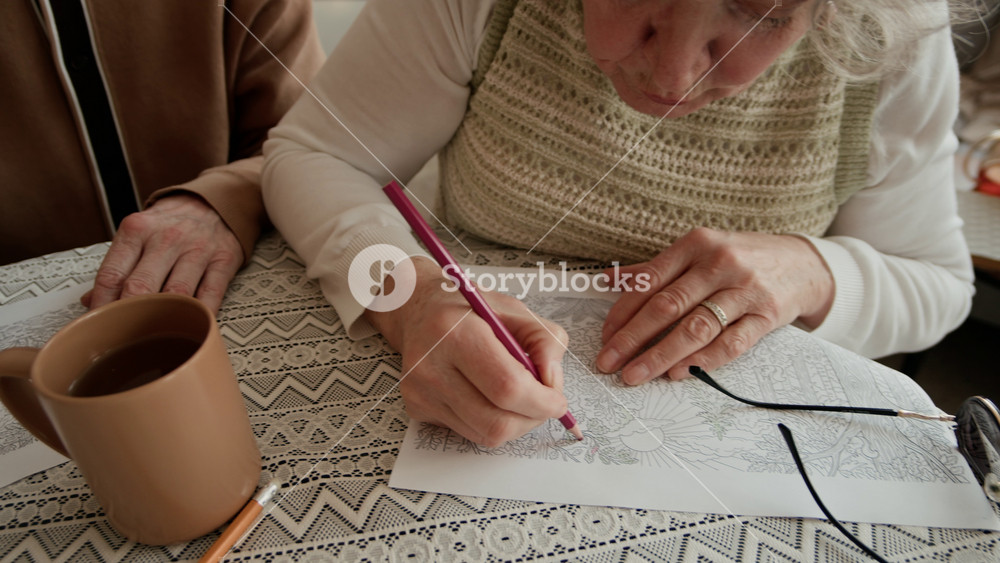 Handheld shot of young Caucasian nurse sitting down at table with group of senior adults coloring with pencils on paper in care home