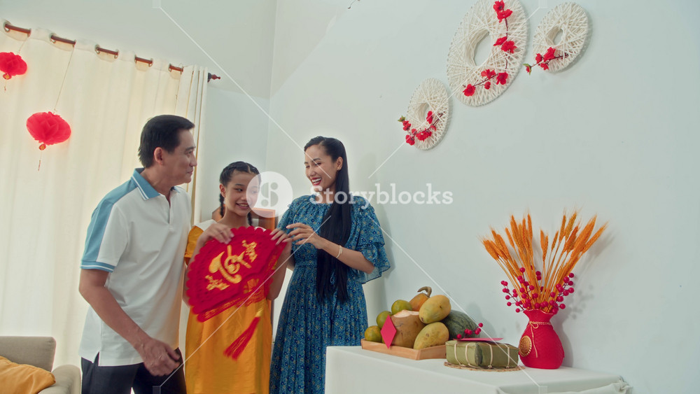 Medium long of parents and their teenage daughter hanging handmade red paper decorations on wall, celebrating Tet at home, text on decoration means Tet