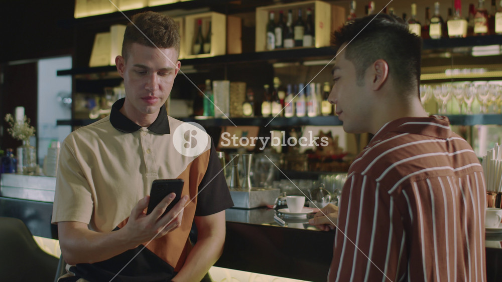 Medium shot of male Caucasian client transferring money to bank account of Asian businessman using application on smartphone in cafe