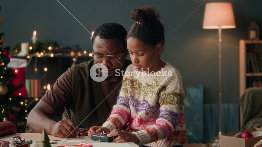 Candid shot of African American man with little daughter sitting on lap writing letter to Santa together or expressing warm wishes on Christmas card in cozy living room