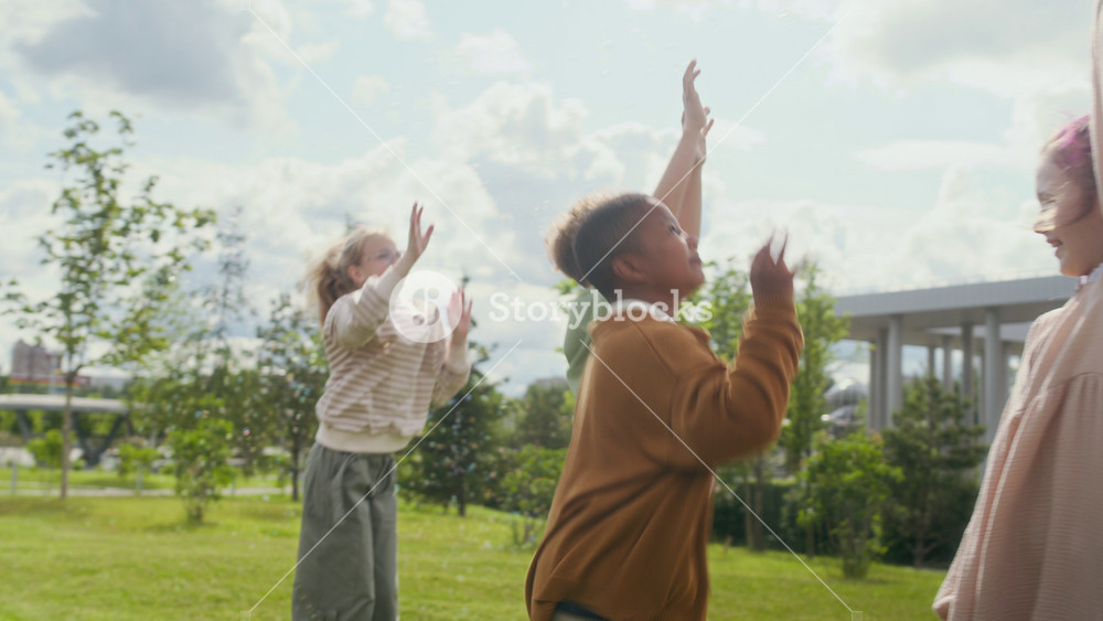 Two ecstatic intercultural boys and blond girl jumping on green lawn in park while trying to catch stream of soap bubbles flying out of plastic toy gun and floating in the air