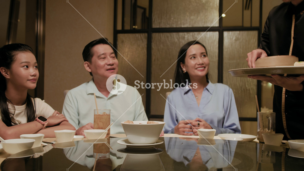 Wide shot of Asian family of daughter, father and mother looking at waiter serving their table with various dishes in restaurant