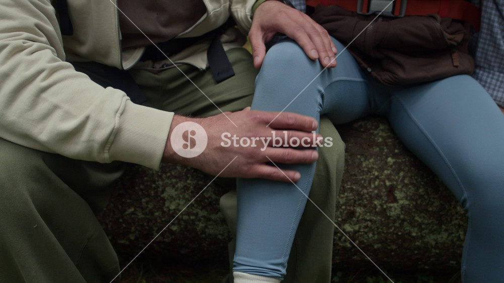 Handheld shot of young male hiker checking injured leg of his girlfriend while sitting on fallen log in summer forest after outdoor accident