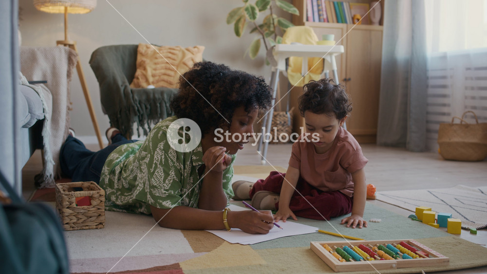 Handheld shot of young African American mother and cute little son drawing on paper with pencils while resting on floor in cozy living room
