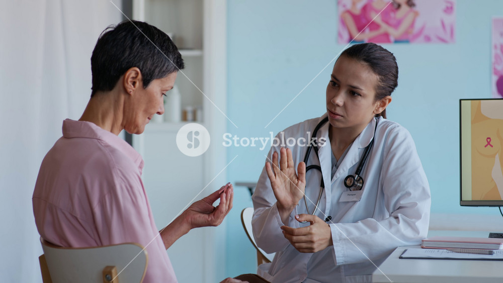 Medium shot of female Caucasian doctor in white coat with stethoscope handing pink ribbon breast cancer awareness pin to mature patient during consultation in clinic office