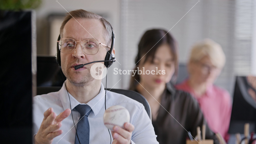 Stab shot of adult male Caucasian operator holding baseball consulting clients via headset while coworking with colleagues in call center office