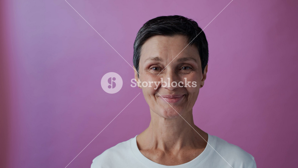 Tilt up portrait of female middle-aged breast cancer survivor wearing white t-shirt and pink ribbon smiling at camera against pink background, copy space